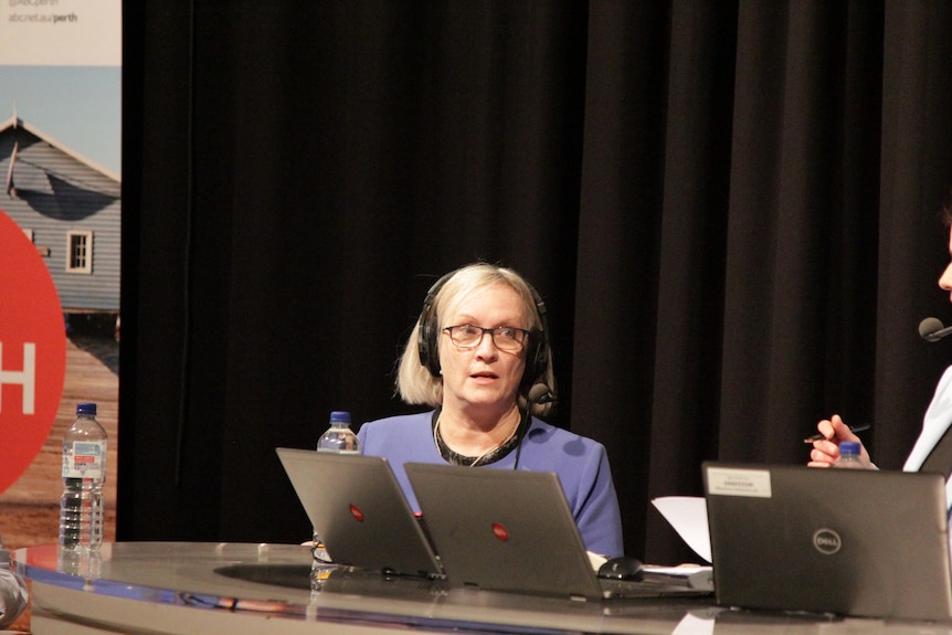 A woman with grey hair and glasses sits in front of a laptop, wearing headphones.