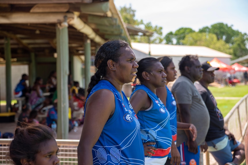 Trois femmes autochtones portant des maillots de football bleus se tiennent sur la touche en regardant un match de football.