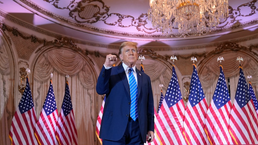 Donald Trump holds his fist up in the air looking defiant with US flags around him and a chandelier above him
