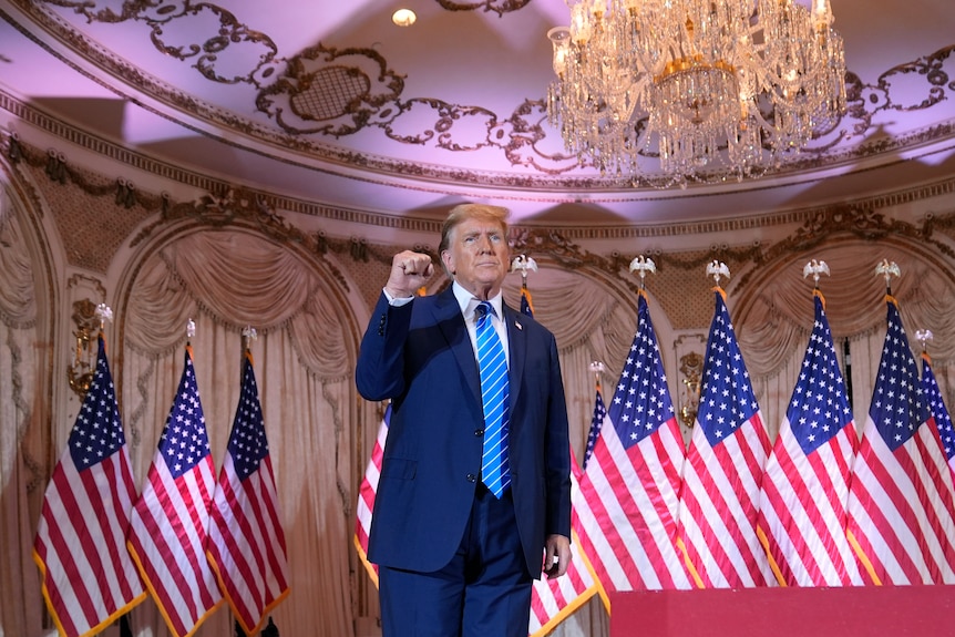 Donald Trump holds his fist up in the air looking defiant with US flags around him and a chandelier above him