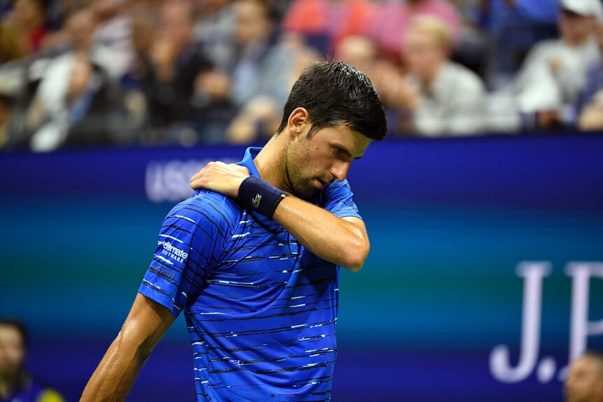 A male tennis player holds his right shoulder as he looks towards the ground.