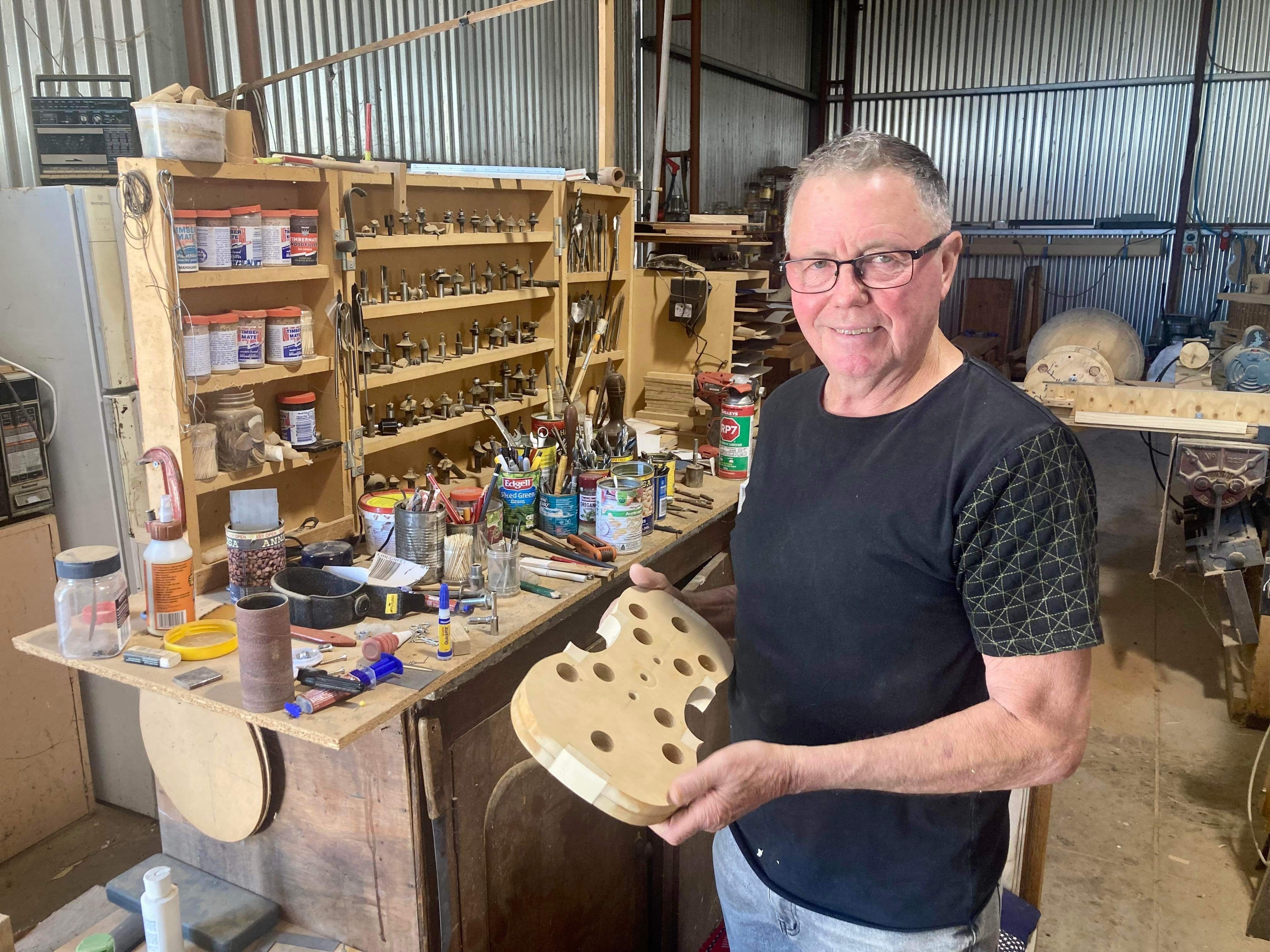 A man with short grey hair wears a dark tshirt, holds a wooden violin frame in a tin shed
