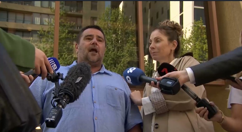 A man in a blue shirt and a woman in a trench coat speak to the media