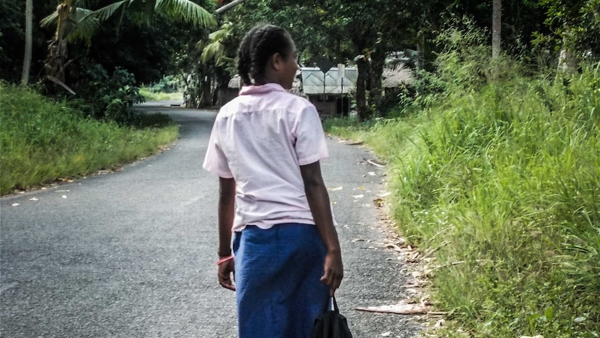 A school school wearing her uniform walks to school on a road surrounded by green brush