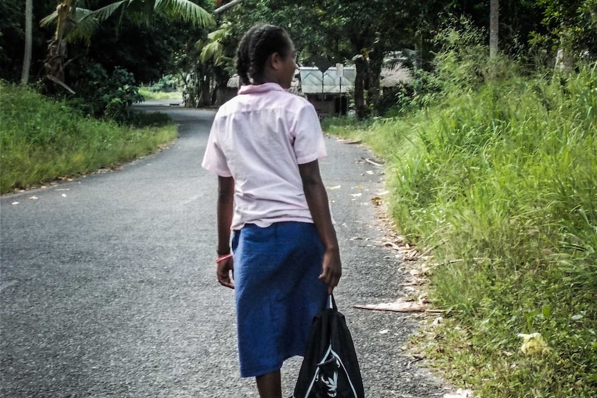 A school school wearing her uniform walks to school on a road surrounded by green brush