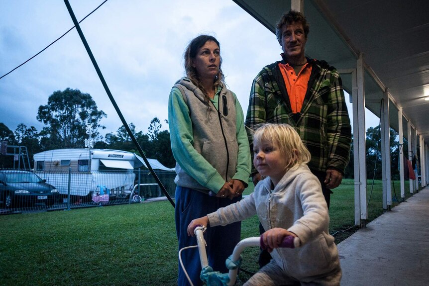 Jodie and Michael stand behind their daughter riding a tricycle in a caravan park.