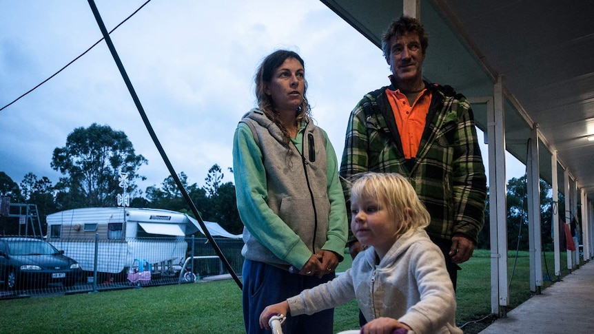 Jodie and Michael stand behind their daughter riding a tricycle in a caravan park.