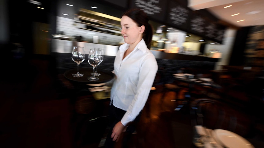 A waitress in a white shirt carries a tray of glasses in a Canberra restaurant.