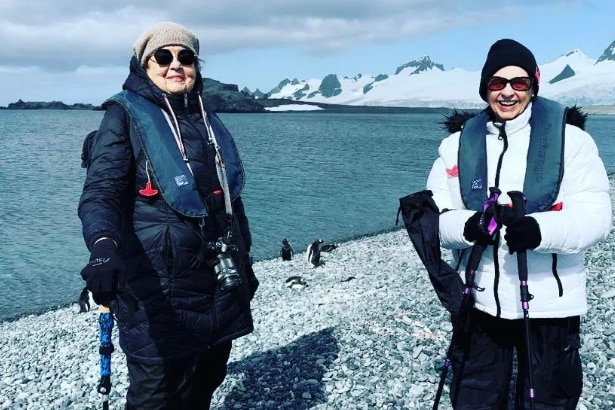 Two ladies standing on a snow covered beach with penguins in the background 