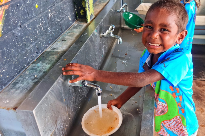 A young boy in a school uniform smiles at the camera by a bubbler.