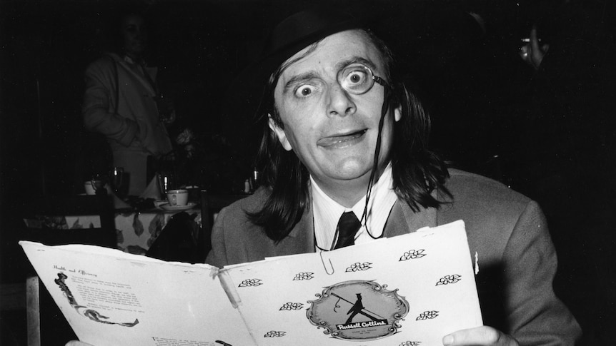 A black and white photo of Barry Humphries wearing a monocle while reading a menu