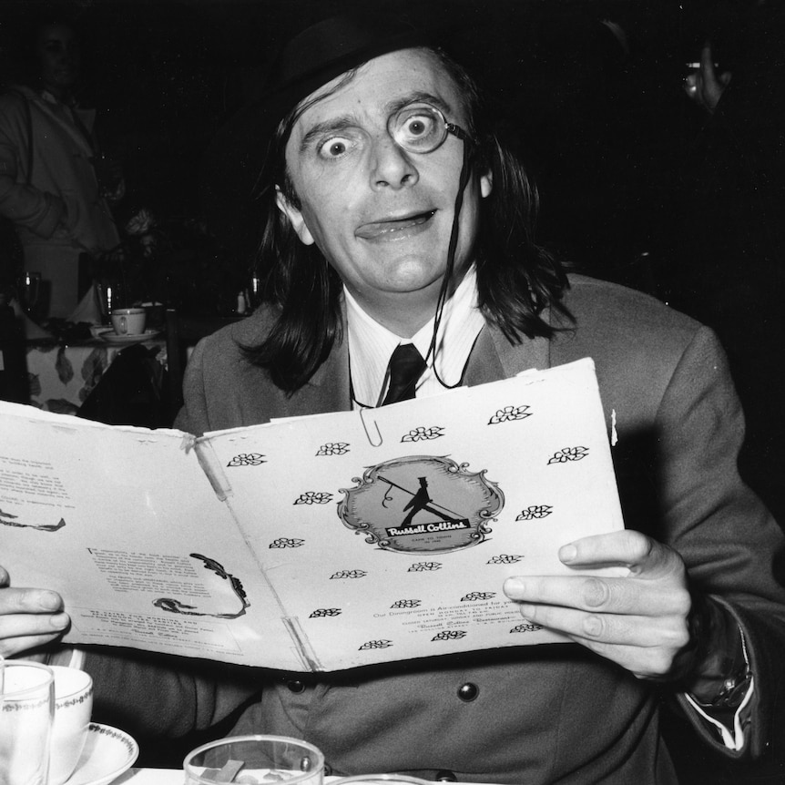 A black and white photo of Barry Humphries wearing a monocle while reading a menu