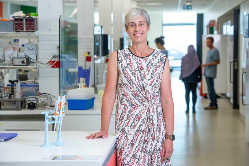 A woman in a colourful patterned dress leans on a laboratory bench and smiles.