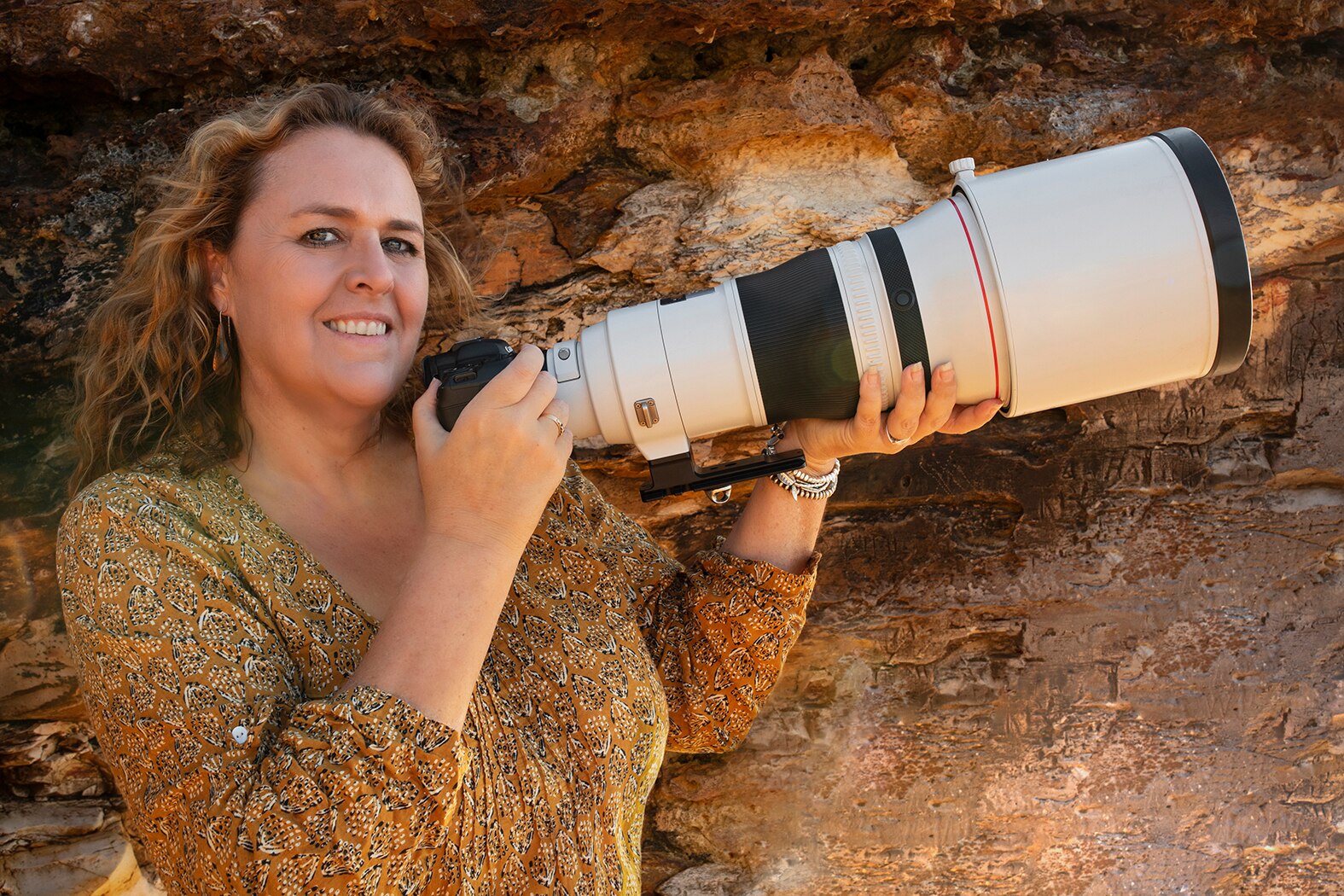 Woman holding large camera with red dirt rock in the background.