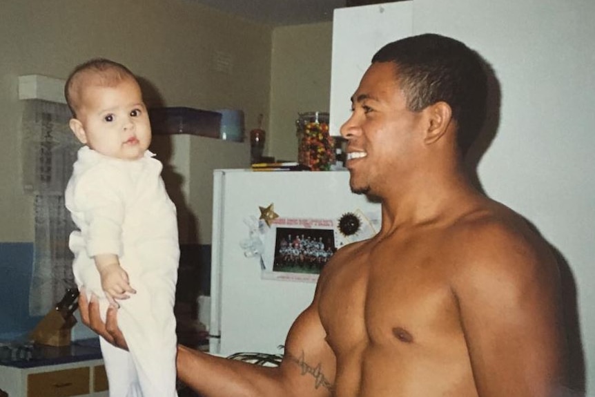 A shirtless man holds a baby up with one hand