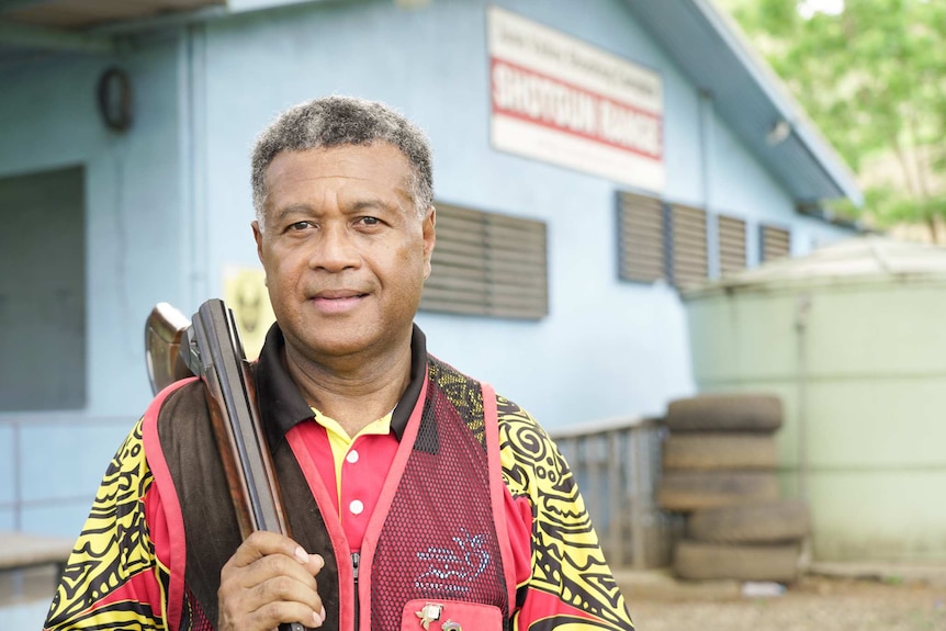 A man with dark skin and wearing a red and yellow shirt smiles with a gun on his shoulder.