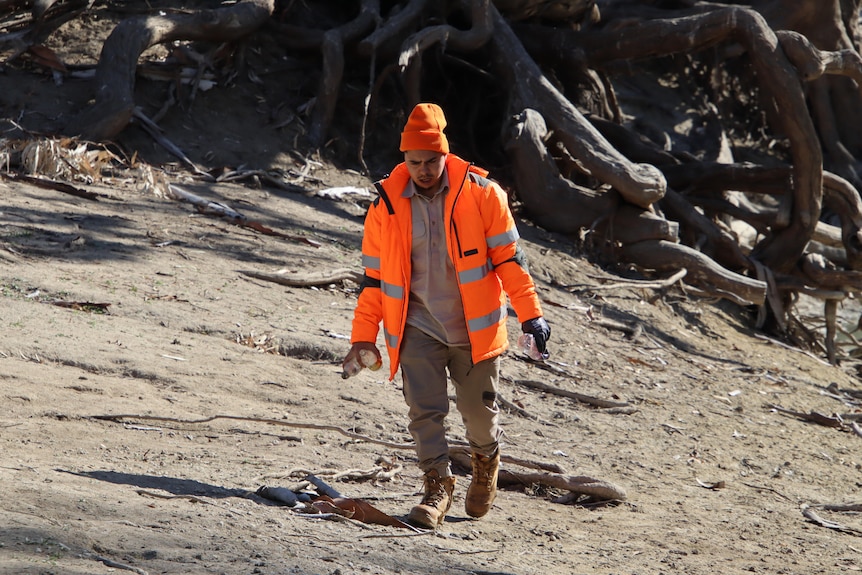 a man in a fluro orange coat and beanie carrying plastic bottles along the river bank