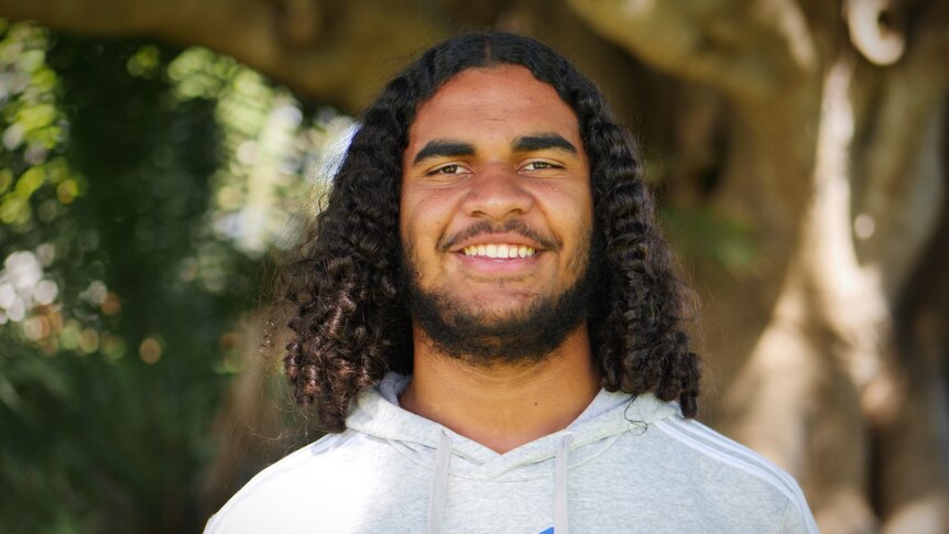 A young Indigenous man with curly shoulder-length hair in a grey hoodie in front of a fig tree