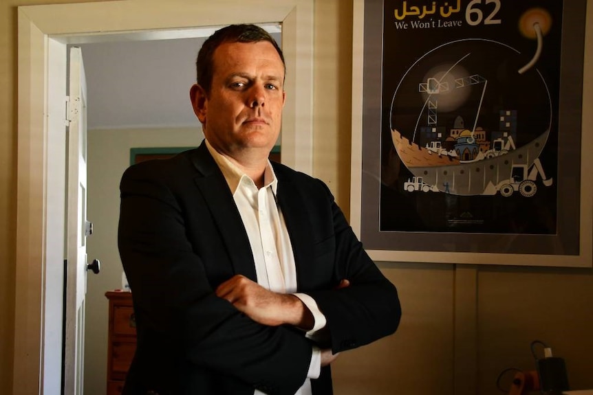 A dark-haired man wearing a suit stands in an office with his arms crossed, looking stern.