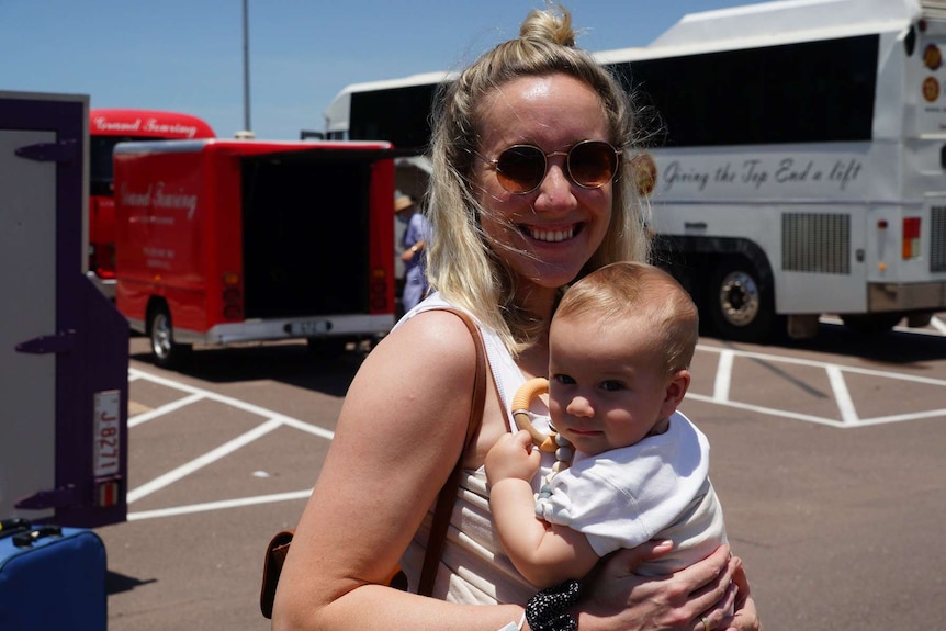 A woman in sunglasses smiles while holding a small baby in a carpark.