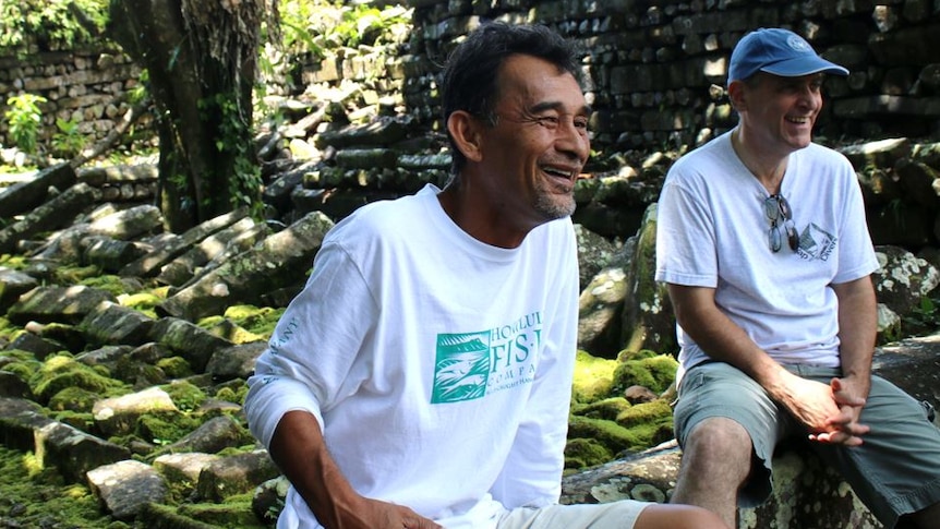 A man wearing a white shirt and shorts sits on a mossy rock wall smiling 