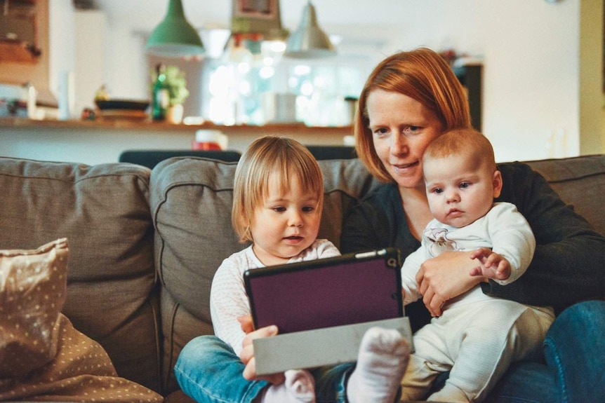 A woman with red hair looks at a tablet with her two children.