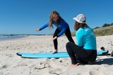 A child stands on a surfboard on the beach as a woman kneels next to her for a story about surf therapy for autistic children