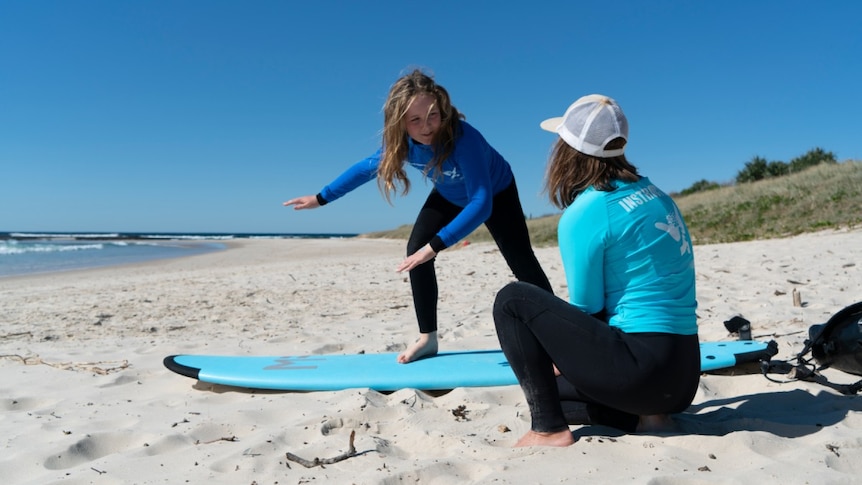 A child stands on a surfboard on the beach as a woman kneels next to her for a story about surf therapy for autistic children