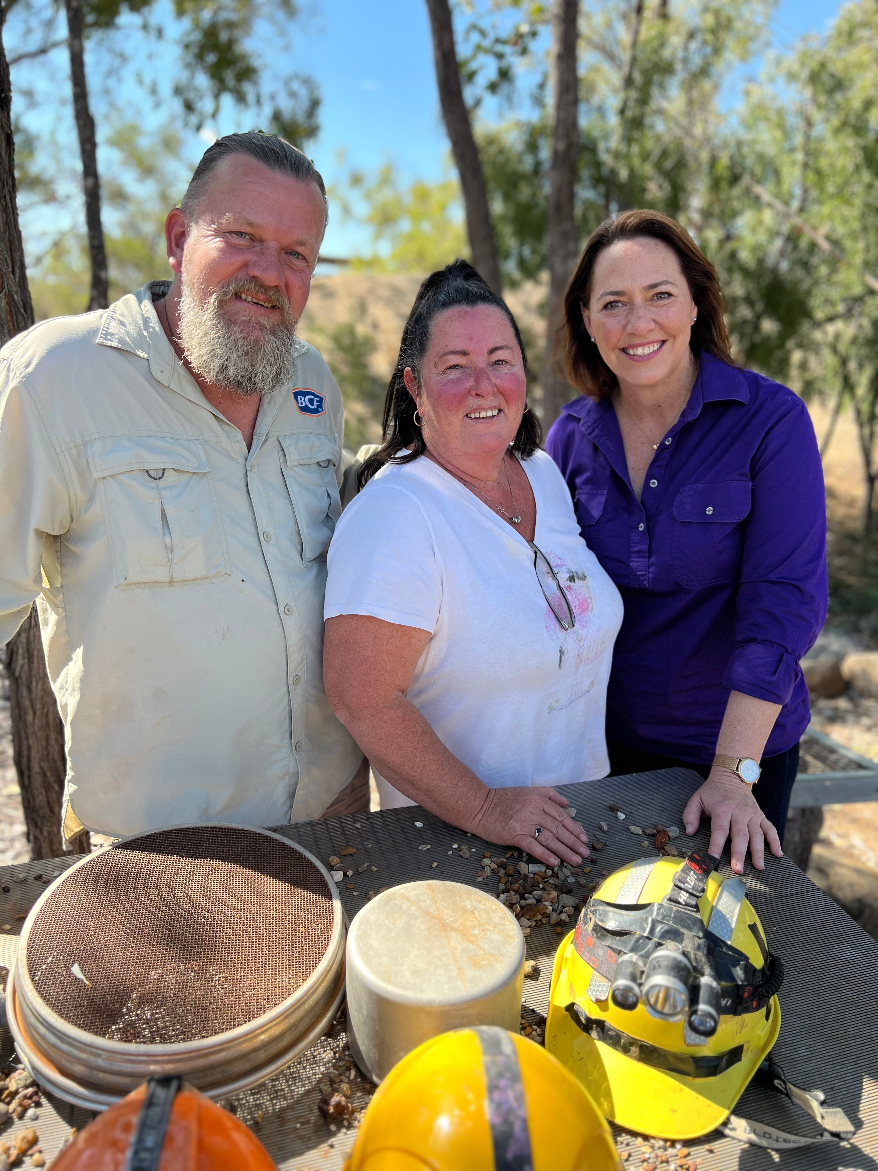 A man with beard and two women smile with scrub behind them
