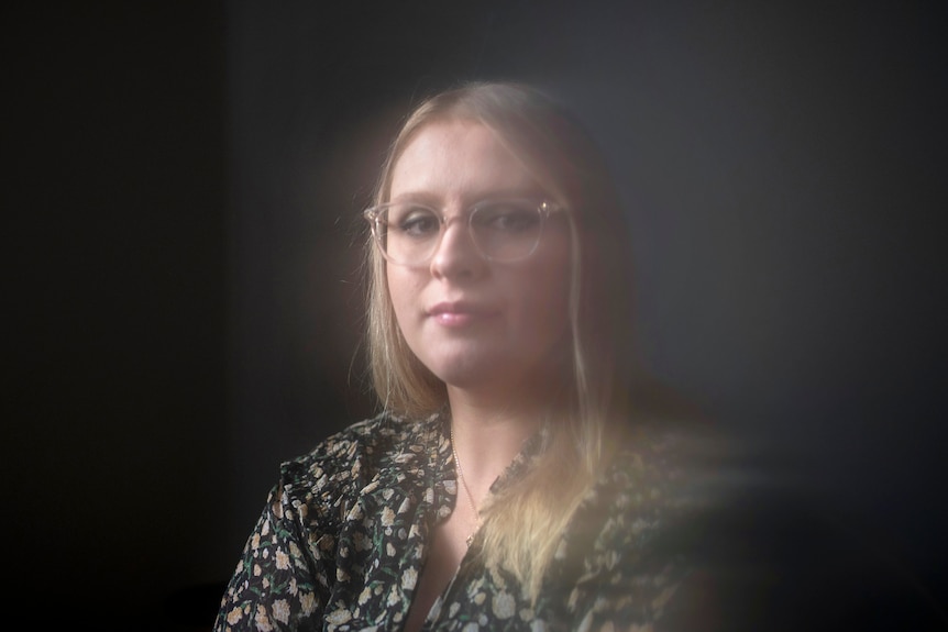 A blonde woman wearing a floral blouse and glasses looks into the camera in soft, cloudy light and dark background.