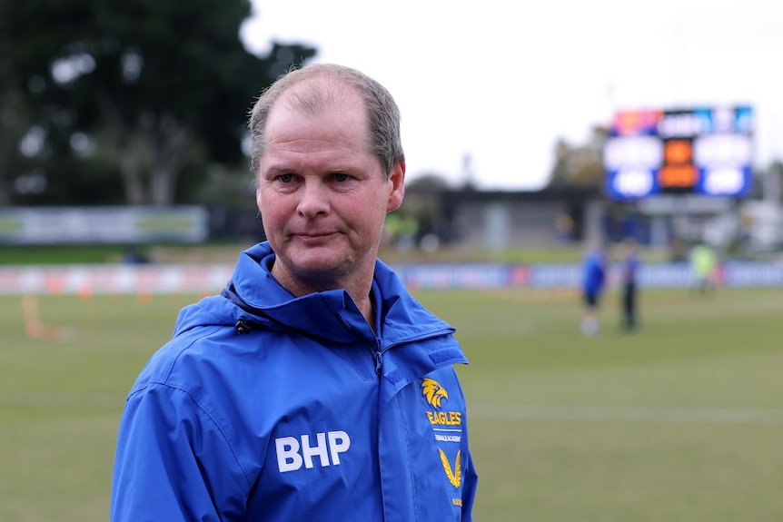 A balding man wearing a blue jacket at a football ground. 