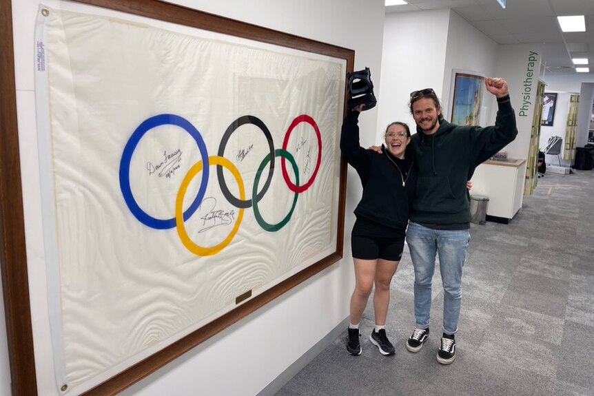 A man and a woman stand next to a framed olympic ring flag. The woman holds a brace in the air. Both are excited