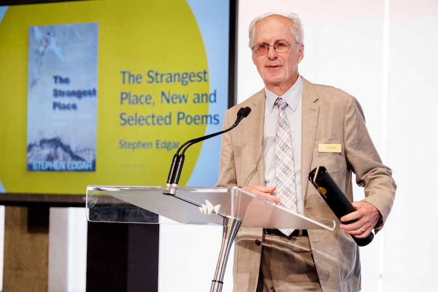 A 70-year-old balding man with glasses stands at a podium in front of a slideshow showing a book cover