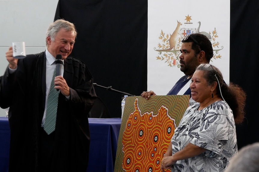 A man in a suit holding a mug and two people holding an Indigenous painting