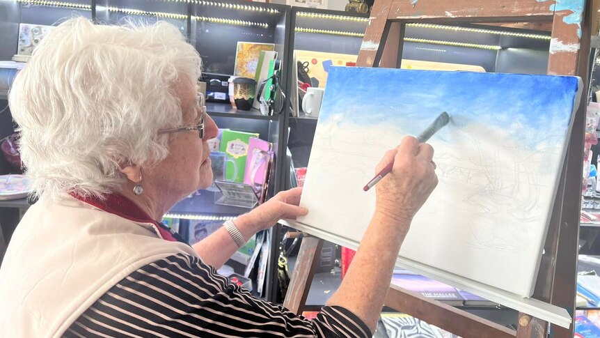 An older woman holds a paint brush up to a canvas