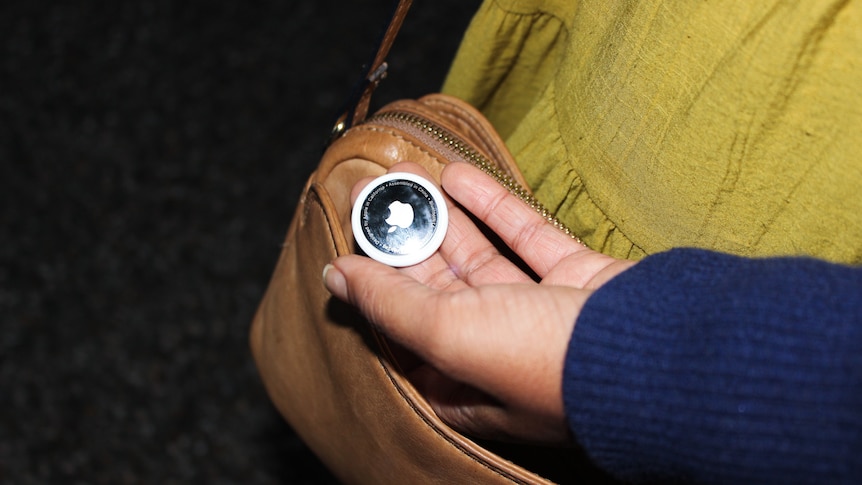 A woman holds an Airtag in her hand near her bag.