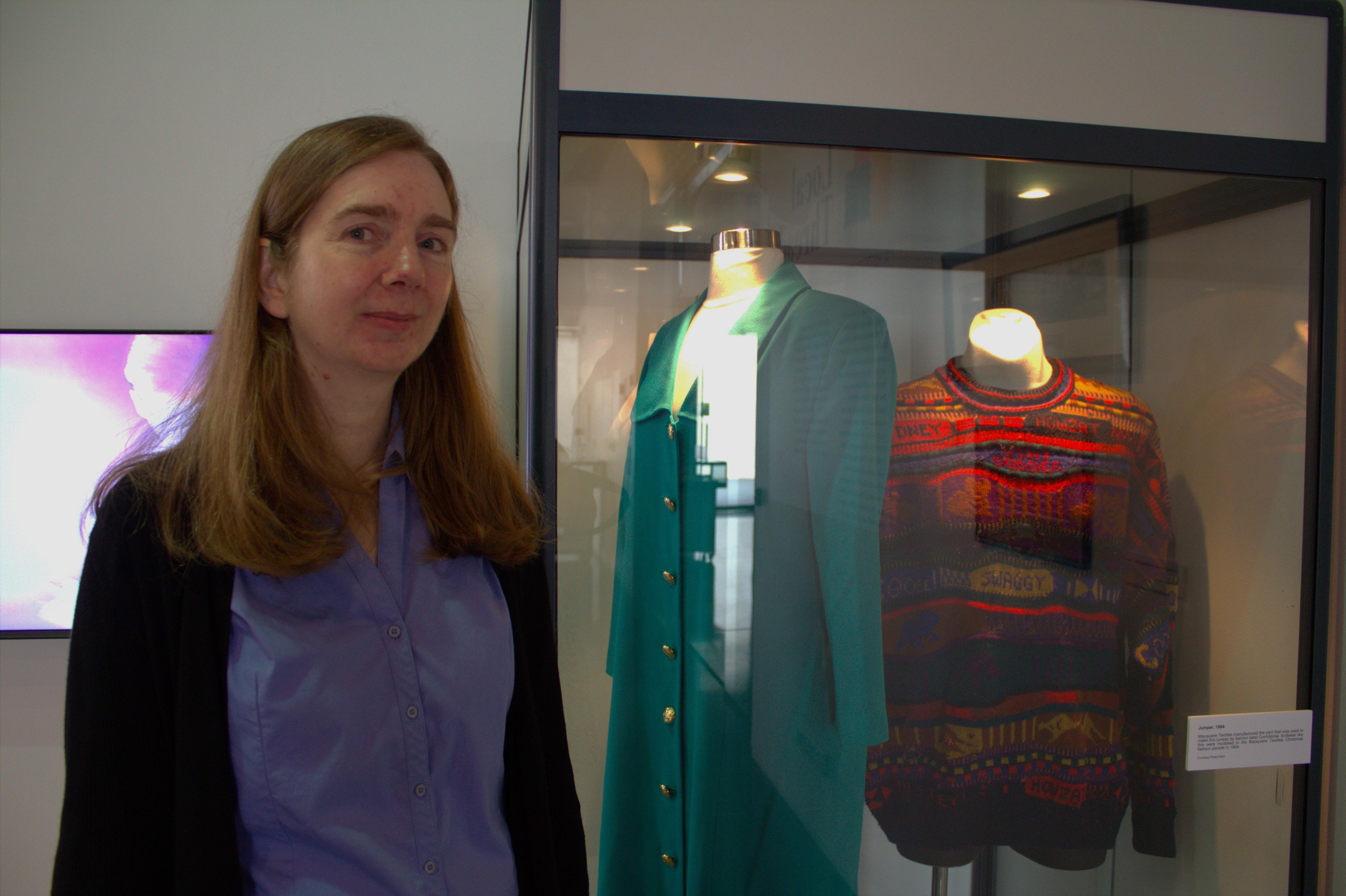 Smiling Sandra with brown hair stands next to a glass case exhibiting clothing on mannequins. 