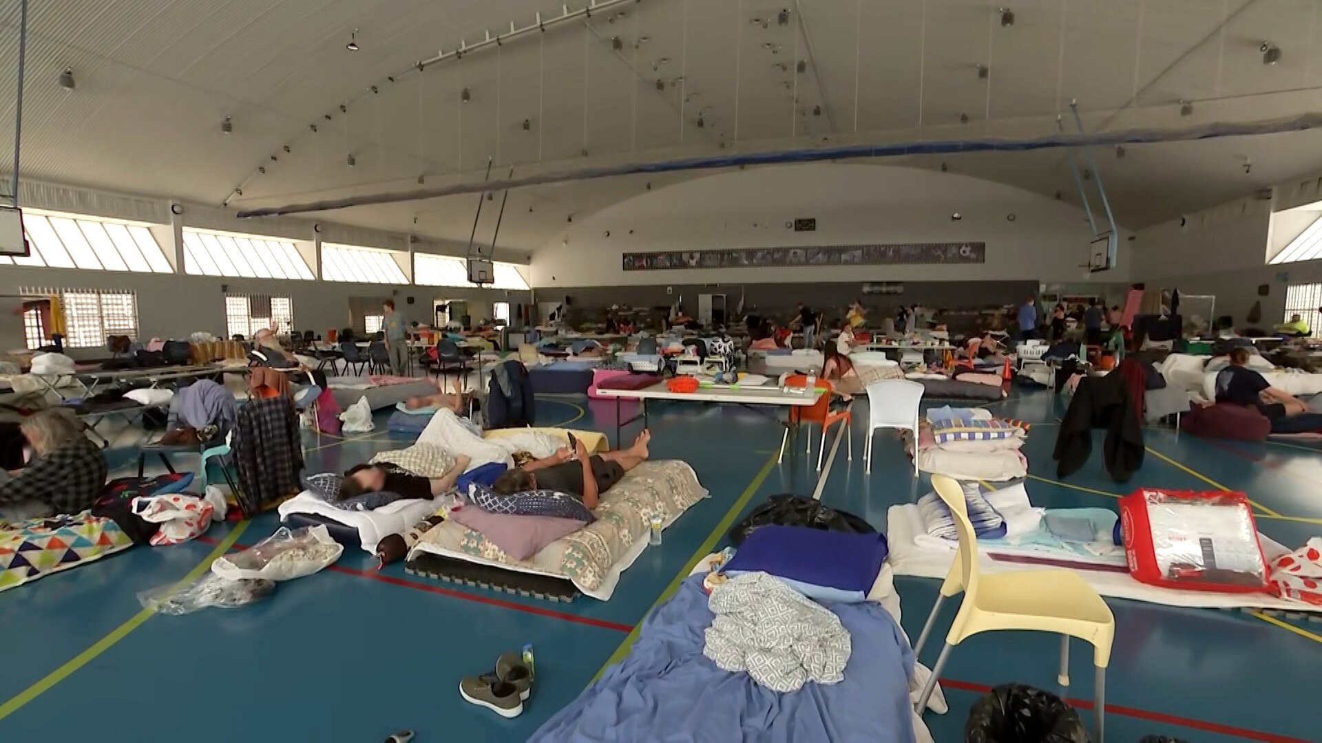An indoor basketball room filled with people, bedding, chairs and belongings