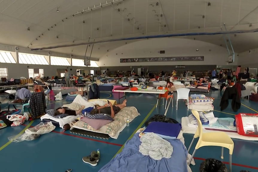 An indoor basketball room filled with people, bedding, chairs and belongings