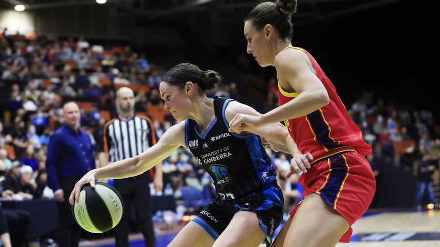 A female basketballer bouncing a ball with her arm out to a defender in a red uniform