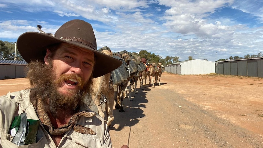 A bearded man with a brown hat and a number of camels walks along a dirt road on a sunny day