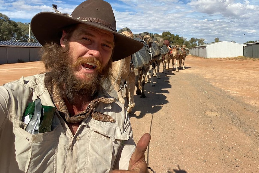 A bearded man with a brown hat and a number of camels walks along a dirt road on a sunny day