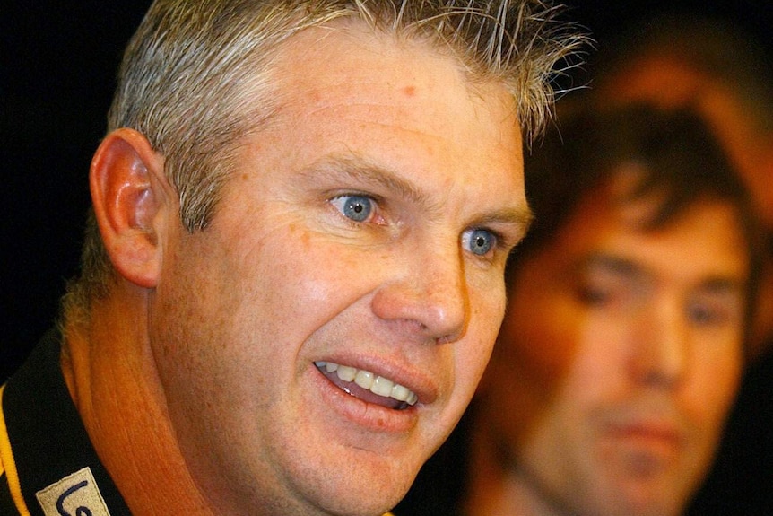 Danny Frawley, wearing a black and yellow Richmond shirt, speaks at a press conference.