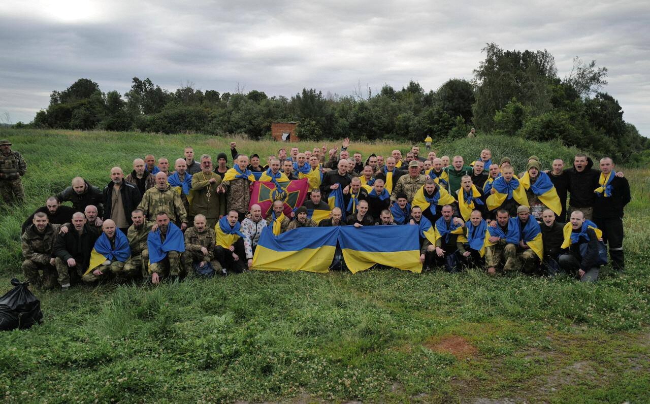 Ukrainian prisoners of war hold a blue and yellow Ukraine flag as they sit in a group and pose for a picture on a grass clearing