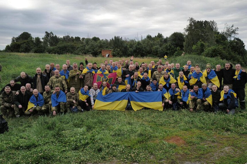 Ukrainian prisoners of war hold a blue and yellow Ukraine flag as they sit in a group and pose for a picture on a grass clearing