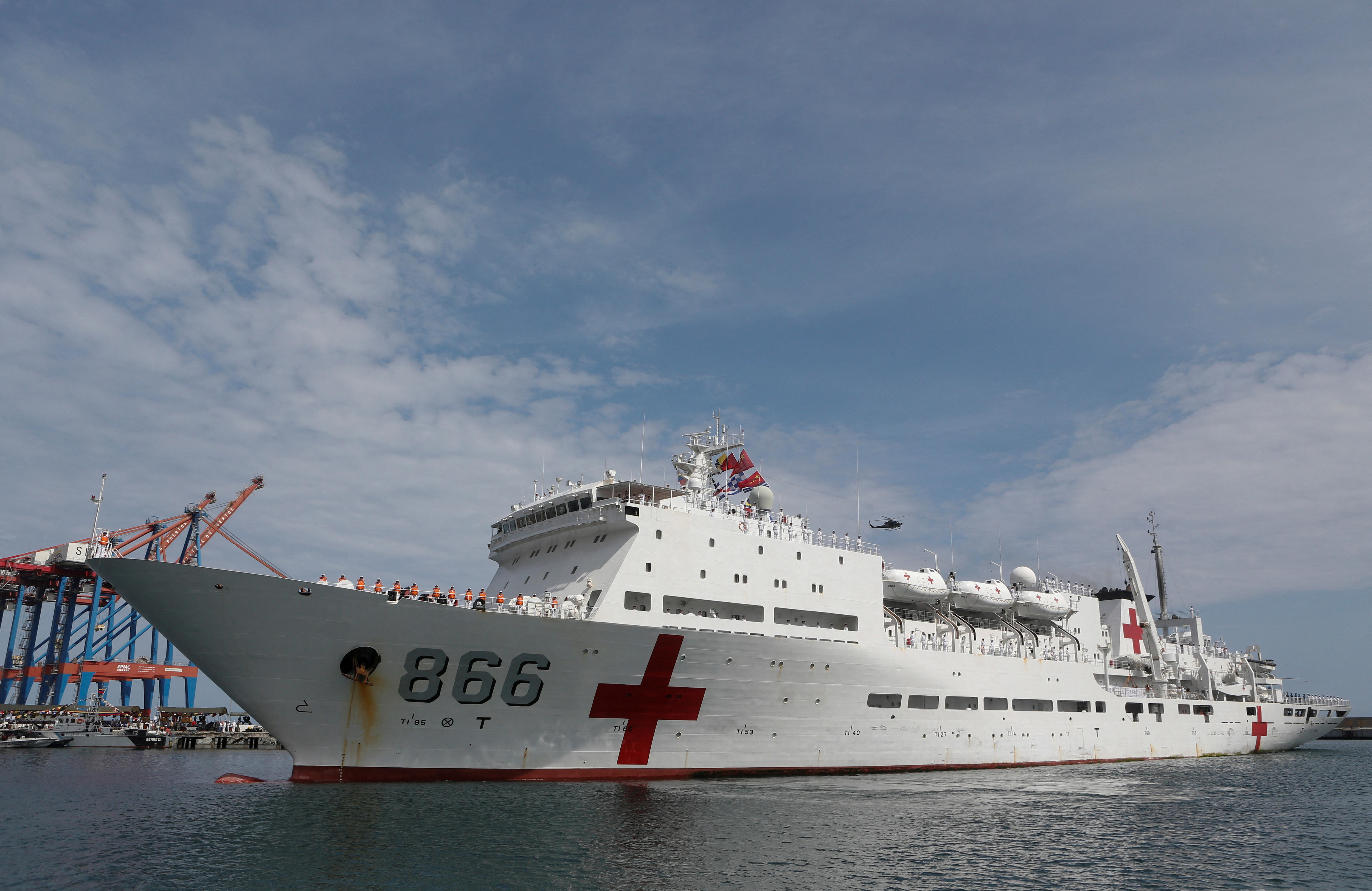 A white China's People's Liberation Army Navy hospital ship with a red cross painted near its bow sits in flat water at a port.