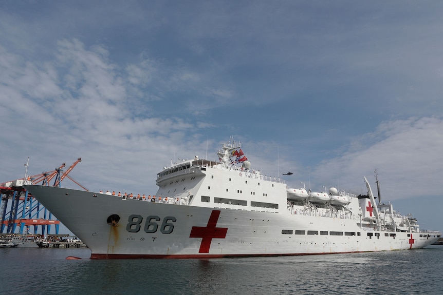 A white China's People's Liberation Army Navy hospital ship with a red cross painted near its bow sits in flat water at a port.
