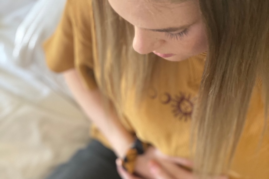 A teenage girl with mousey brown hair looks down and holds her stomach as she sits on a bed.