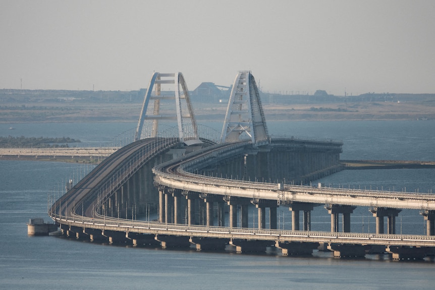 A large, double-span bridge curves across a blue-grey body of water in the daytime.