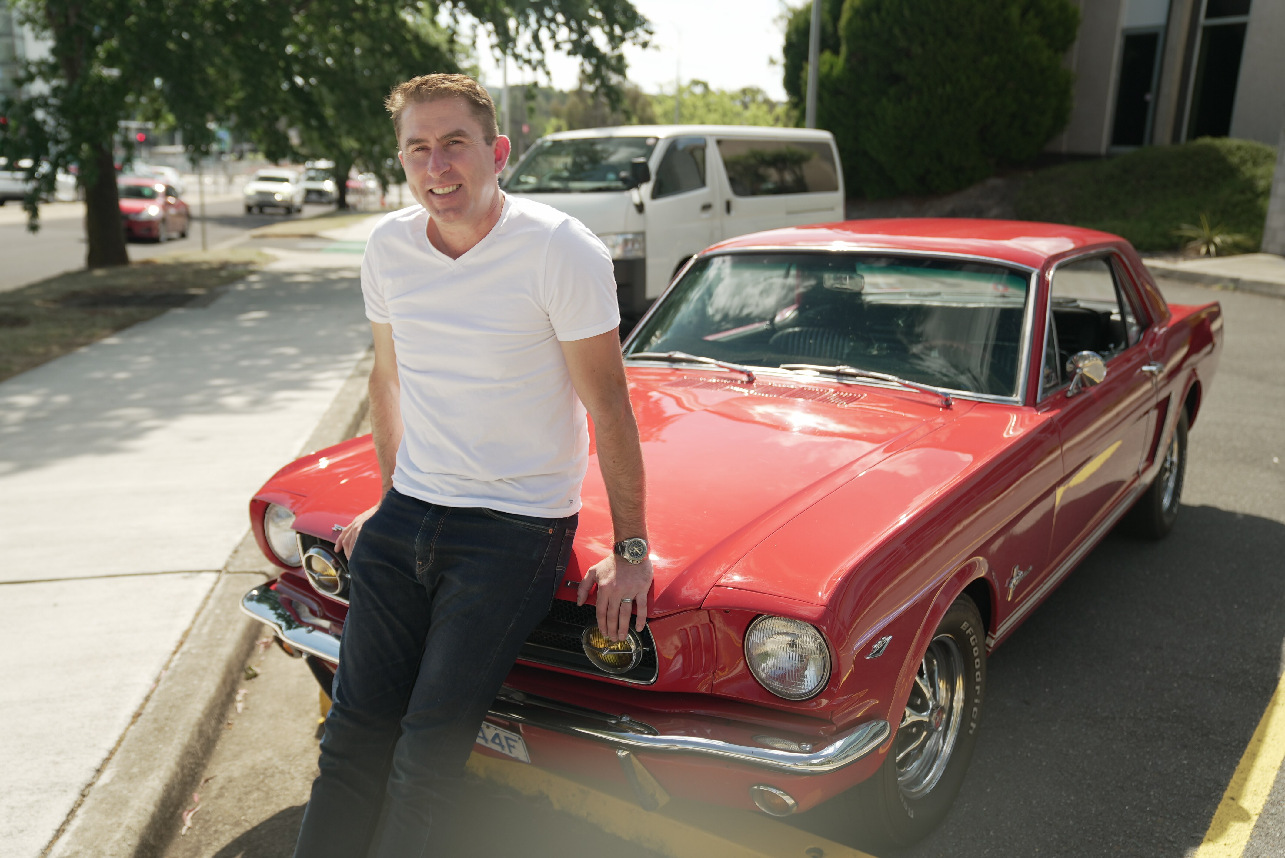 Nick leans against his red Mustang car, wearing a white shirt and jeans.